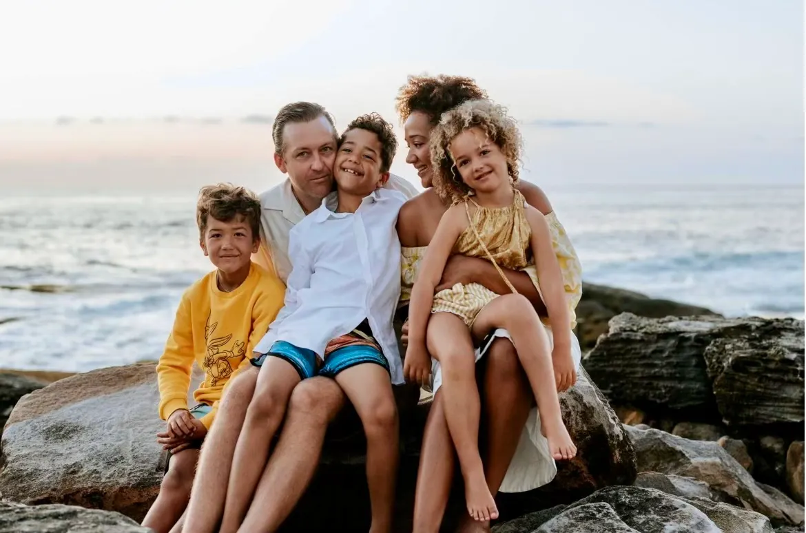 Sam Spry with his family, sitting together on sandstone rocks beside the ocean.